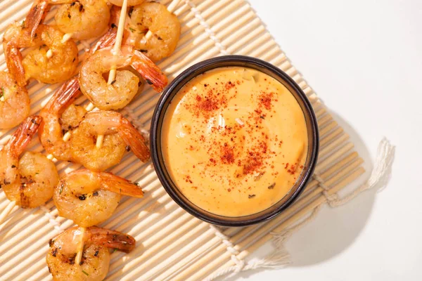 Vista superior de sabrosos langostinos en brochetas con salsa sobre estera de bambú sobre fondo blanco - foto de stock