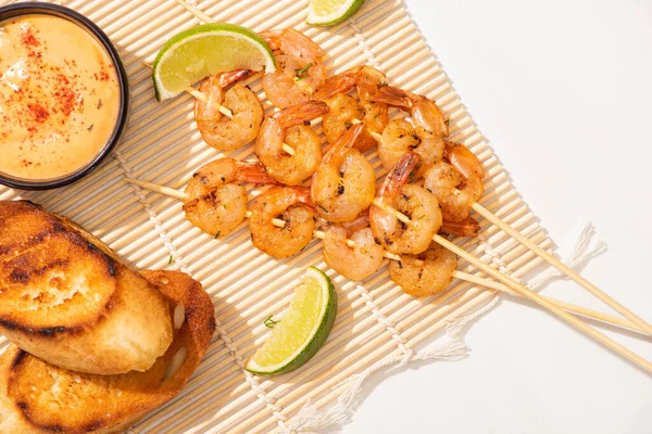Top view of prawns on skewers with grilled bread, lime and sauce on bamboo mat on white background — Stock Photo