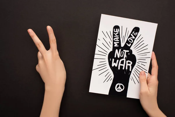 Cropped view of woman showing peace sign and holding white paper with hand drawing and make love not war lettering on black background — Stock Photo