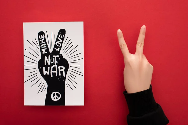 Cropped view of woman showing peace sign near drawing with make love not war lettering and peace sign on red background — Stock Photo