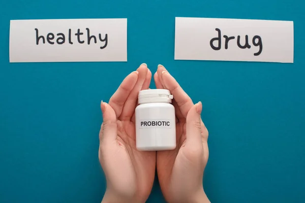 Cropped view of woman holding white probiotic container near cards with healthy and drug words on blue background — Stock Photo