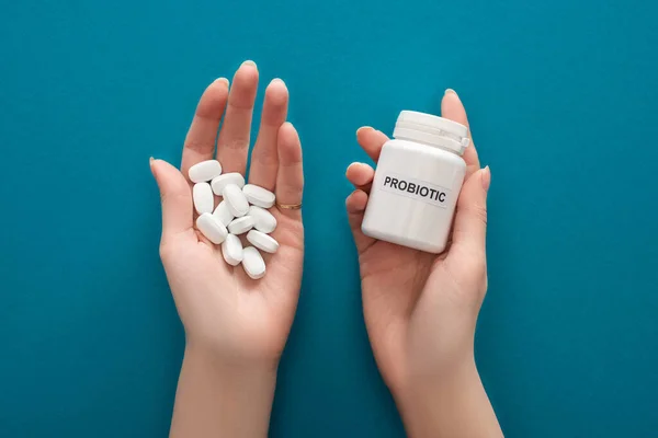 Vista recortada de la mujer sosteniendo contenedor probiótico blanco y pastillas en las manos sobre fondo azul - foto de stock