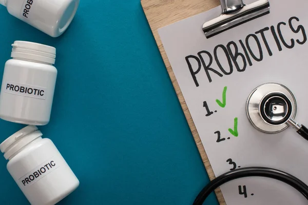 Top view of clipboard with probiotics empty list with stethoscope near containers with pills on blue background — Stock Photo