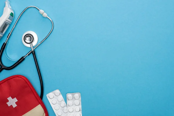 Vue du dessus des pilules en plaquettes thermoformées, trousse de premiers soins, thermomètre auriculaire et stéthoscope sur fond bleu — Photo de stock