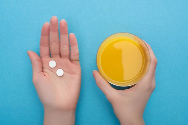 Vista parcial de la mujer sosteniendo pastillas y jugo de naranja sobre fondo azul — Stock Photo