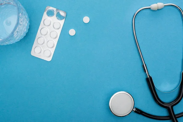 Вид таблеток в блистерной упаковке, стакан воды и стетоскоп на синем фоне — стоковое фото