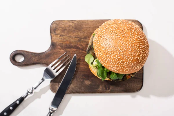 Вид на вкусный веганский бургер на деревянной доске с столовыми приборами на белом фоне — стоковое фото