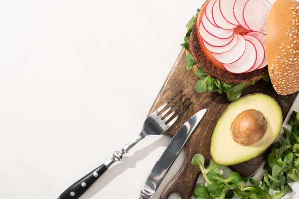 Draufsicht auf leckeren veganen Burger mit Rettich, Avocado und Gemüse auf Holzschneidebrett mit Besteck auf weißem Hintergrund — Stockfoto