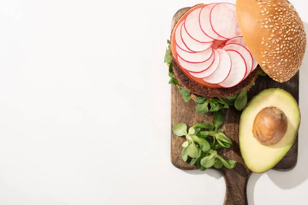 Вид зверху на смачний веганський бургер з редькою, авокадо та зеленню на дерев'яній обробній дошці на білому тлі — стокове фото