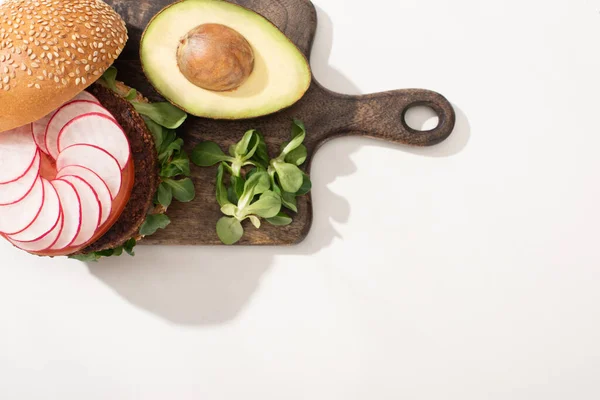 Вид на вкусный веганский бургер с редиской, авокадо и зеленью на деревянной доске на белом фоне — стоковое фото
