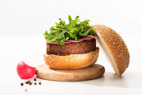 Delicioso hambúrguer vegan com rabanete e rúcula em tábua de madeira perto de pimenta preta no fundo branco — Fotografia de Stock