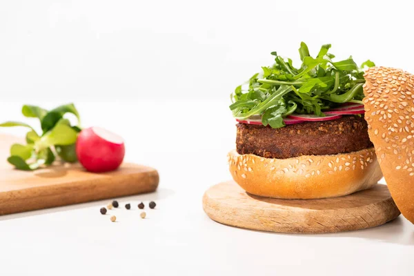 Selektiver Schwerpunkt leckerer veganer Burger mit Rettich und Rucola auf Holzbrett in der Nähe von schwarzem Pfeffer auf weißem Hintergrund — Stockfoto