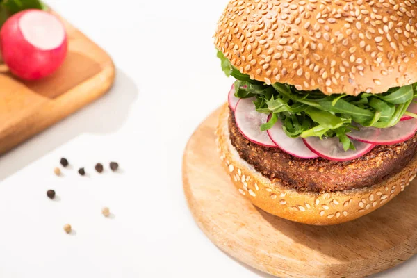 Enfoque selectivo de deliciosa hamburguesa vegana con rábano y rúcula en tablero de madera cerca de pimienta negra sobre fondo blanco - foto de stock