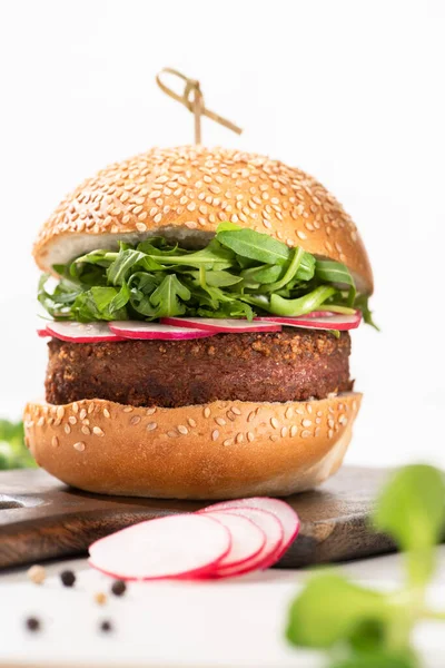 Foco selectivo de deliciosa hamburguesa vegana con rábano y rúcula sobre tabla de madera con pimienta negra sobre fondo blanco - foto de stock