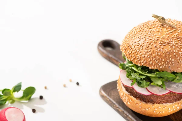 Selektiver Schwerpunkt leckerer veganer Burger mit Rettich und Rucola auf Holzbrett mit schwarzem Pfeffer auf weißem Hintergrund — Stock Photo