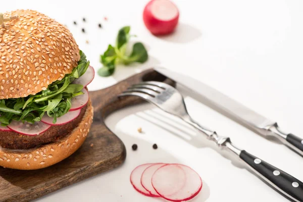 Foco seletivo de delicioso hambúrguer vegan com rabanete e rúcula em tábua de madeira com pimenta preta perto de garfo e faca no fundo branco — Fotografia de Stock
