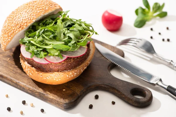 Selective focus of delicious vegan burger with radish and arugula on wooden board with black pepper near fork and knife on white background — Stock Photo