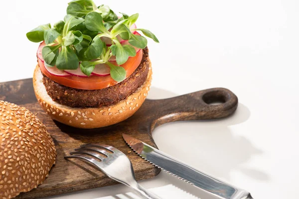 Delicious vegan burger with radish, tomato and microgreens on wooden boar near fork and knife on white background — Stock Photo