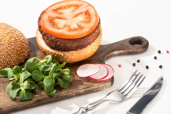 Selective focus of delicious vegan burger with radish, tomato and microgreens on wooden boar with black pepper near fork and knife on white background — Stock Photo
