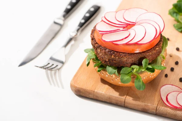 Selective focus of delicious vegan burger with radish, tomato and microgreens on wooden boar with black pepper near fork and knife on white background — Stock Photo