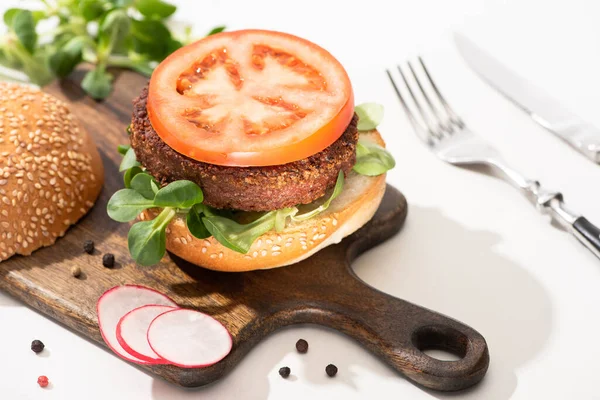 Selective focus of delicious vegan burger with radish, tomato and microgreens on wooden boar with black pepper near fork and knife on white background — Stock Photo