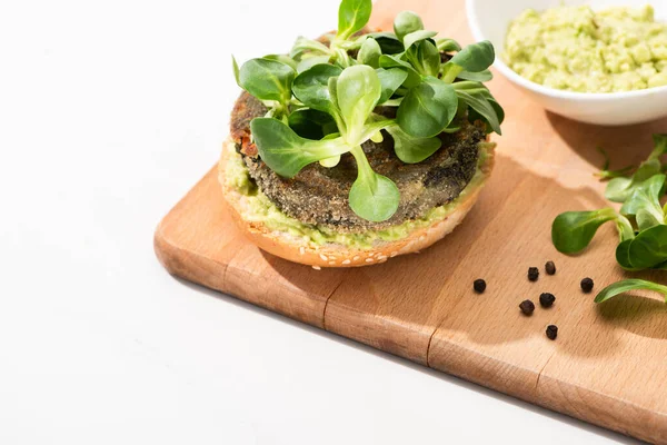 Selective focus of delicious green vegan burger with microgreens and mashed avocado on wooden boar with black pepper on white background — Stock Photo