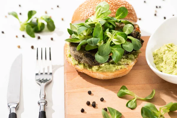 Delicious green vegan burger with microgreens and mashed avocado on wooden boar with black pepper near fork and knife on white background — Stock Photo