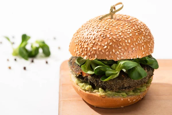 Selective focus of delicious green vegan burger with microgreens and mashed avocado on wooden boar with black pepper on white background — Stock Photo