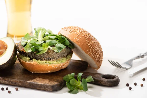 Selective focus of delicious green vegan burger with microgreens, avocado, black pepper on wooden cutting board near fork and oil on white background — Stock Photo