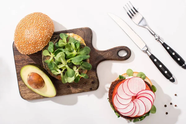 Draufsicht auf vegane Burger mit Mikrogemüse, Avocado, Rettich auf Holzschneidebrett auf weißem Hintergrund mit Besteck — Stockfoto
