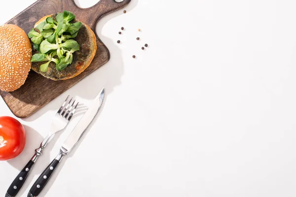 Vue de dessus du hamburger végétalien avec microgreens sur planche de bois près de tomate et couverts sur fond blanc — Photo de stock