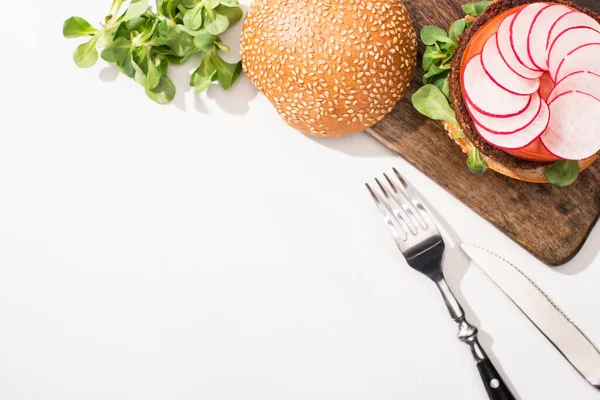 Vue de dessus du hamburger végétalien avec microgreens, radis sur planche à découper en bois près des couverts sur fond blanc — Photo de stock