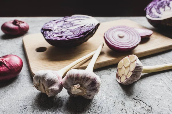 Selective focus of red cabbage, onion and garlic on wooden cutting board on grey concrete surface isolated on black — Stock Photo