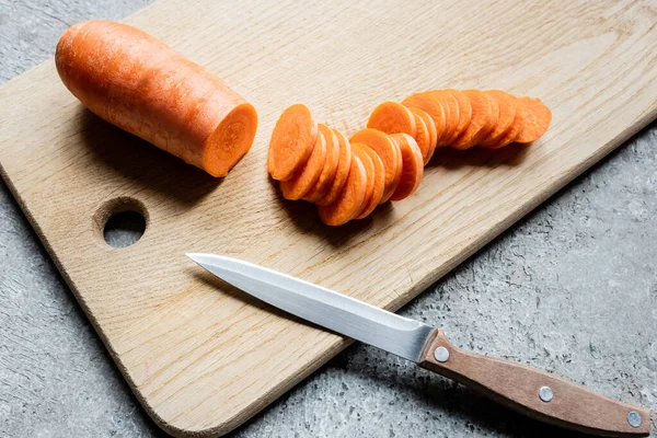 Fresh ripe carrot slices on wooden cutting board with knife on concrete surface — Stock Photo