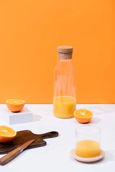 Zumo de naranja fresco en vidrio y botella cerca de fruta cortada en tabla de cortar de madera con cuchillo en la superficie blanca aislada en naranja - foto de stock