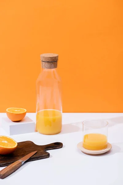 Fresh orange juice in glass and bottle near cut fruit on wooden cutting board with knife on white surface isolated on orange — Stock Photo