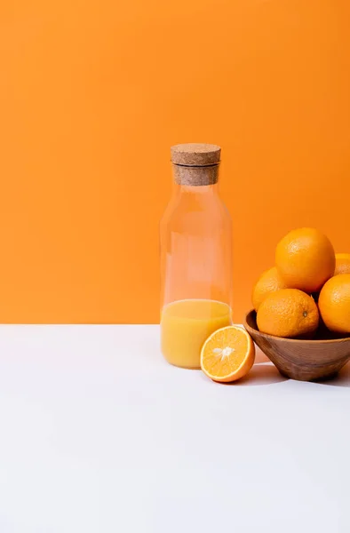 Suco de laranja fresco em garrafa de vidro perto de laranjas em tigela na superfície branca isolada em laranja — Fotografia de Stock