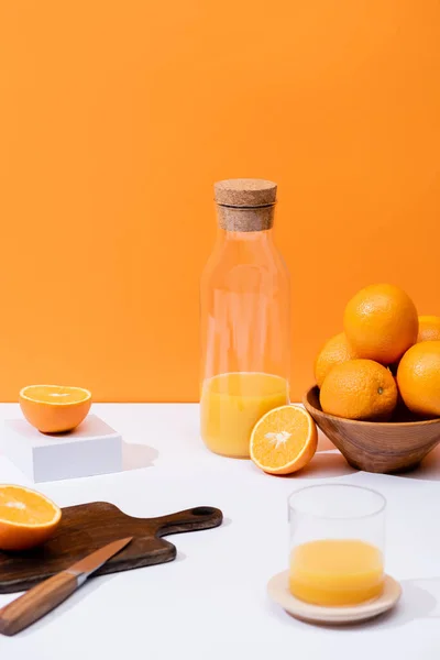 Zumo de naranja fresco en vidrio y botella cerca de naranjas en tazón, tabla de cortar de madera con cuchillo en la superficie blanca aislada en naranja - foto de stock