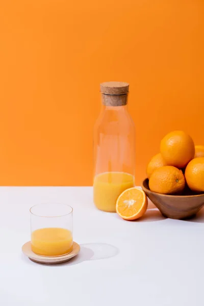 Jus d'orange frais en verre et bouteille près des oranges dans un bol sur surface blanche isolé sur orange — Photo de stock