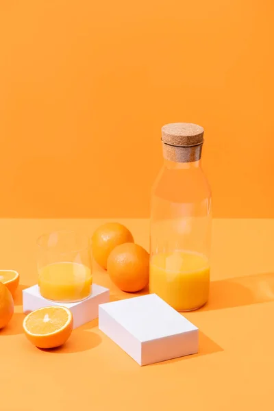 Jus d'orange frais en verre et bouteille près des oranges mûres et des cubes blancs isolés sur orange — Photo de stock