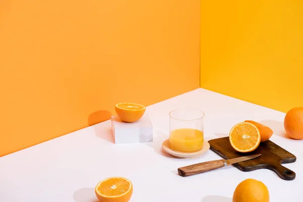 Fresh orange juice in glass near ripe oranges, wooden cutting board with knife on white surface on orange background — Stock Photo