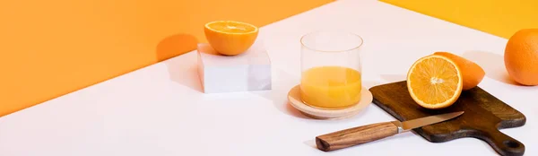 Fresh orange juice in glass near ripe oranges, wooden cutting board with knife on white surface on orange background, panoramic shot — Stock Photo