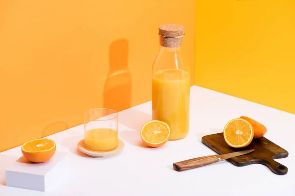 Frischer Orangensaft in Glas und Flasche in der Nähe reifer Orangen, Holzschneidebrett mit Messer auf weißer Oberfläche auf orangefarbenem Hintergrund — Stockfoto