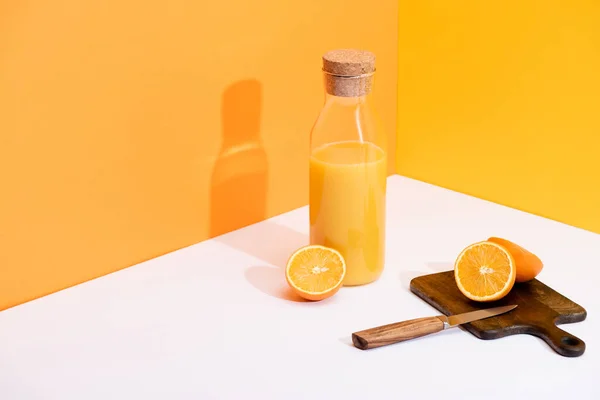 Suco de laranja fresco em garrafa de vidro perto de laranjas maduras, placa de corte de madeira com faca na superfície branca no fundo laranja — Fotografia de Stock