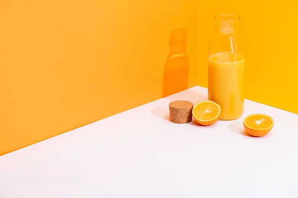 Свіжий апельсиновий сік у скляній пляшці поблизу стиглих апельсинів та пробки на білій поверхні на помаранчевому фоні — стокове фото