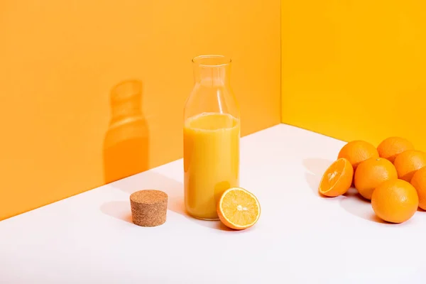 Suco de laranja fresco em garrafa de vidro perto de laranjas maduras e cortiça na superfície branca no fundo laranja — Fotografia de Stock