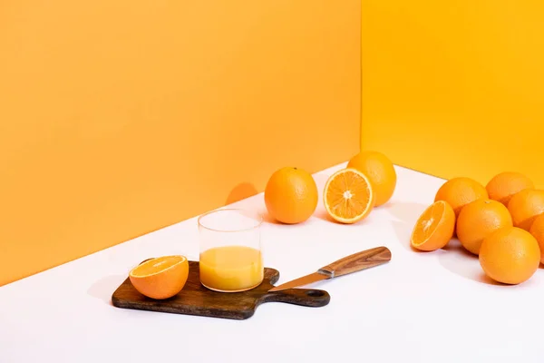 Fresh orange juice in glass on wooden cutting board with knife near ripe oranges on white surface on orange background — Stock Photo