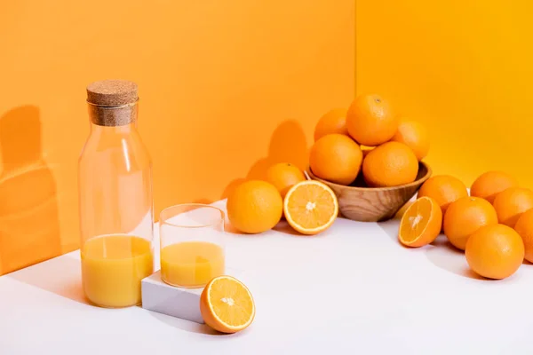 Suco de laranja fresco em vidro e garrafa perto de laranjas maduras em tigela na superfície branca no fundo laranja — Fotografia de Stock