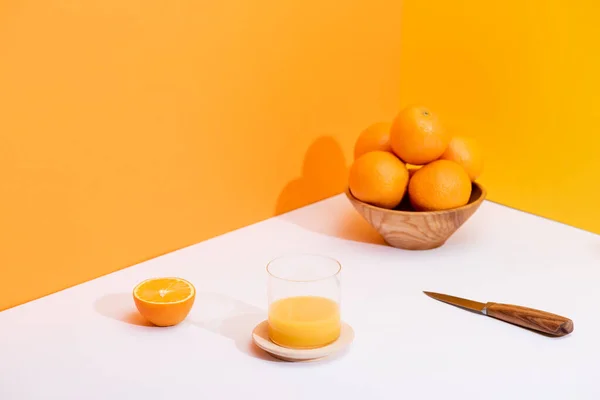 Zumo de naranja fresco en vaso cerca de naranjas maduras en tazón y cuchillo sobre superficie blanca sobre fondo naranja - foto de stock