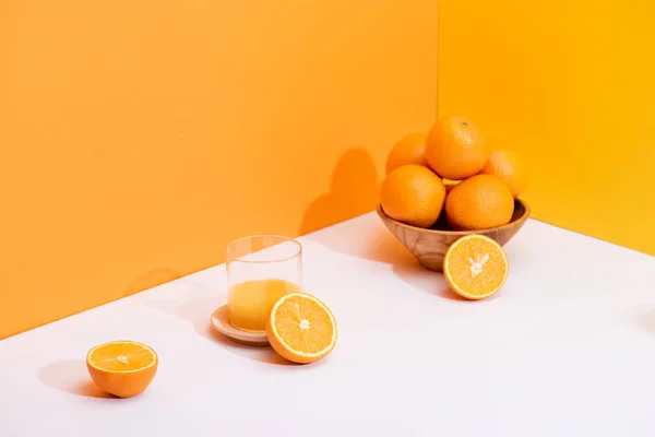 Suco de laranja fresco em vidro perto de laranjas maduras em tigela na superfície branca no fundo laranja — Fotografia de Stock